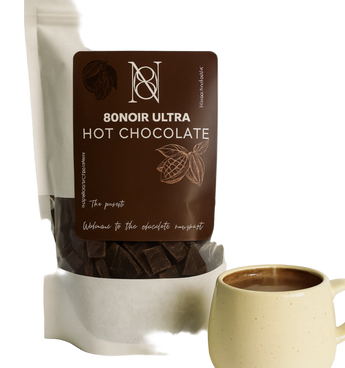 A stand-up pouch of 8onoir Ultra Hot Chocolate with a large dark brown label, shown next to a light beige ceramic mug filled with hot chocolate on a clean white background.
