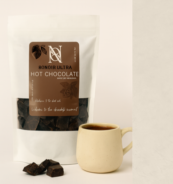 A white stand-up pouch of 8NOIR ULTRA hot chocolate with a large brown label, shown next to a cream ceramic mug filled with hot chocolate and a few dark chocolate pieces on a light surface.