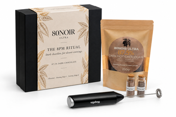 A curated chocolate and coffee ritual set on a white background, including a black and beige 80Noir Ultra gift box, a brown stand-up pouch of 80Noir Ultra dark hot chocolate, two small glass vials, a black Keplin handheld frother, and a metal whisk attachment.