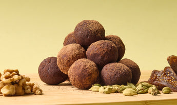 A pyramid of chocolate truffles dusted with cocoa powder sits on a wooden board, surrounded by whole walnuts, green cardamom pods, and dried figs against a soft yellow-green background.
