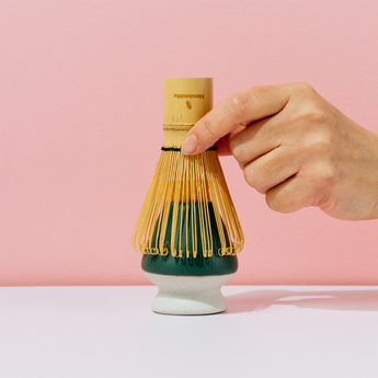 Hand holding a bamboo whisk over a green matcha bowl against a pink background