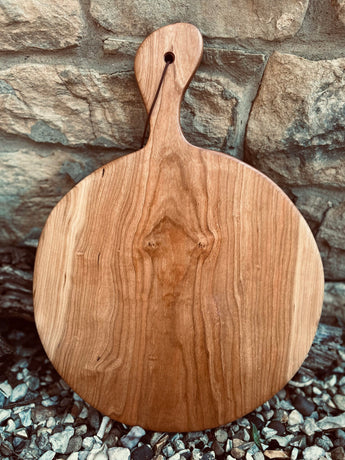 A round wooden serving board with a single handle and a hanging hole, leaning against a stone wall on gravel.