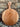A round wooden serving board with a single handle and a hanging hole, leaning against a stone wall on gravel.
