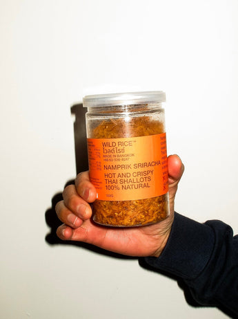 A hand holding a clear plastic jar of Namprik Sriracha hot and crispy Thai shallots, featuring a bright orange label and visible dried red-orange spices inside, set against a plain off-white background.
