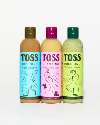 Three bottles of Toss Dressing arranged side by side on a white background. The bottles contain vinaigrette, salad chick, and green goddess dressings, with colorful labels in blue, pink, and green, and matching caps.