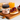 A jar of Crwst Almond Honey Butter sits on a wooden board next to an open slice of toast topped with honey and red berries, with a glass of orange juice and more toast in the softly lit background.