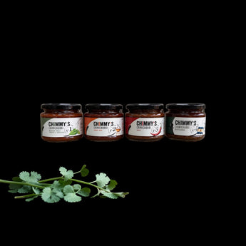 Four jars of Chimmy's hot sauces arranged on a rustic wooden block over a wooden table, with cilantro, peppercorns, and chili peppers as props against a black background.