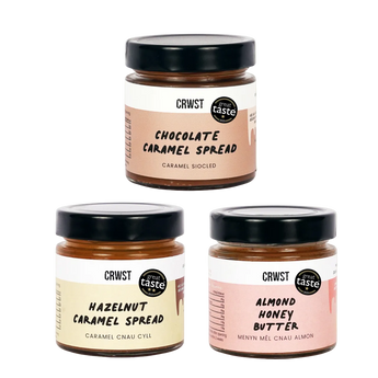 Three jars of CRWST caramel spreads arranged in a triangle on a light sage green background: Chocolate Caramel Spread at the top, Hazelnut Caramel Spread at bottom left, and Almond Honey Butter at bottom right. The jars have black lids and labels in peach, cream, and pink tones.