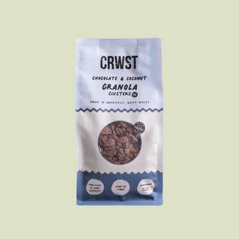 A stand-up pouch of CRWST Chocolate & Coconut Granola Clusters on a solid pale yellow-green background. The packaging is white with blue accents and a circular clear window showing the granola inside.