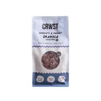 A stand-up pouch of CRWST Chocolate & Coconut Granola Clusters on a solid pale yellow-green background. The packaging is white with blue accents and a circular clear window showing the granola inside.