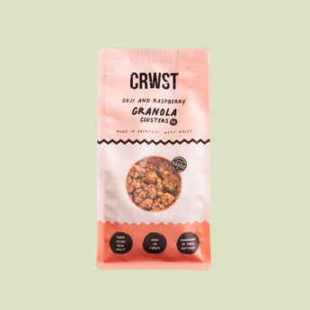 A package of CRWST Granola Clusters in Goji and Raspberry flavor, shown against a solid pale sage green background. The packaging is a soft pink and white bag with black text and a circular window displaying the golden-brown granola inside.