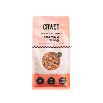 A package of CRWST Granola Clusters in Goji and Raspberry flavor, shown against a solid pale sage green background. The packaging is a soft pink and white bag with black text and a circular window displaying the golden-brown granola inside.