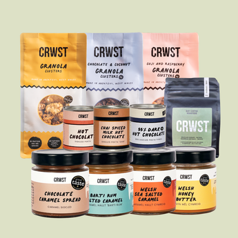 A collection of CRWST branded granola clusters and honey butters arranged on a light sage green background. The assortment includes three large granola cluster bags in yellow, white, and pink, three hot chocolate containers in peach, orange, and dark chocolate, and four honey butters in caramel, spiced caramel, sea salted caramel, and honey.