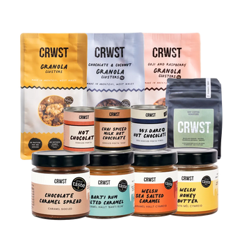 A collection of CRWST branded granola clusters and honey butters arranged on a light sage green background. The assortment includes three large granola cluster bags in yellow, white, and pink, three hot chocolate containers in peach, orange, and dark chocolate, and four honey butters in caramel, spiced caramel, sea salted caramel, and honey.
