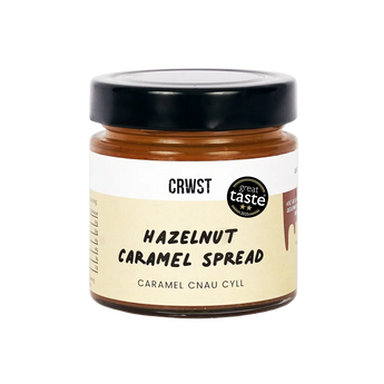 A jar of Crowst Hazelnut Caramel Spread on a solid light sage green background. The product features a black lid, a beige label with black text, and a brown caramel spread visible through the glass jar.