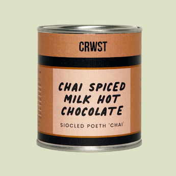 A cylindrical metal can of CRWST Chai Spiced Milk Hot Chocolate, featuring a terracotta-colored label with black text and stripes, set against a solid pale sage green background.