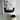 A white stand-up pouch labeled '88NOIR ULTRA HOT CHOCOLATE' with a clear window showing dark chocolate pieces inside, placed on a white surface against a white wall. A small speckled cream mug filled with hot chocolate sits in front.