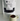 A white stand-up pouch labeled '88NOIR ULTRA HOT CHOCOLATE' with a clear window showing dark chocolate pieces inside, placed on a white surface against a white wall. A small speckled cream mug filled with hot chocolate sits in front.