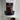 A stand-up pouch of 80noir Ultra Hot Chocolate chocolate, with a large dark brown label and a clear window showing dark chocolate pieces inside. A ceramic mug filled with hot chocolate sits in the foreground against a plain light wall.