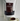 A stand-up pouch of 80noir Ultra Hot Chocolate chocolate, with a large dark brown label and a clear window showing dark chocolate pieces inside. A ceramic mug filled with hot chocolate sits in the foreground against a plain light wall.