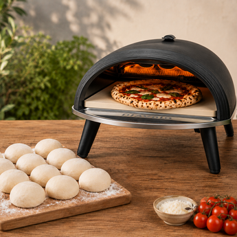 Portable pizza oven with a pizza inside on a wooden table, surrounded by pizza dough and ingredients.