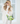 A woman with long blonde hair and sunglasses stands in front of a white window, wearing a cream knit turtleneck and matching pants, while holding a clear tote bag filled with green packaged items and limes.