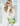 A woman with blonde hair and sunglasses wearing a cream turtleneck sweater and matching pants, holding a clear plastic bag filled with green and white bottles and small green fruits. She is standing in front of a white paneled window.