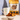 A bowl of granola with cashews and dried fruit sits on a table in front of a branded bag of granola and a white mug on a light surface.