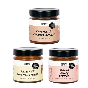 Three jars of CRWST caramel spreads arranged in a triangle on a light sage green background: Chocolate Caramel Spread at the top, Hazelnut Caramel Spread at bottom left, and Almond Honey Butter at bottom right. The jars have black lids and labels in peach, cream, and pink tones.